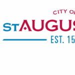 City of St Augustine logo with a crest featuring a fleur de lis, castle, lion, and armored arm with sword Text reads "City of St Augustine Est " in red and blue letters