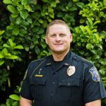 A police officer in a dark uniform stands in front of green leafy bushes, smiling at the camera He has short hair and visible badges on his shirt