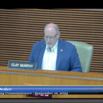 A man in a blue suit and glasses sits at a desk with a nameplate reading "Clay Murphy" during a Board of County Commissioners meeting on September , The screen displays “Call to Order” at the bottom