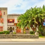 A historic two story villa with red accents, arched windows, palm trees in front, and a colorful neon sign reading "Villa Zorayda" on a stone wall beside the sidewalk