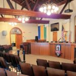 An empty council chamber with rows of wooden seats, a wooden podium, a curved desk, flags, and a blue curtain behind the council area under a high ceiling with exposed beams and chandeliers