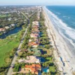 Aerial view of a coastal neighborhood with colorful houses lining a narrow strip of land, bordered by a golf course on the left and a sandy beach with ocean waves on the right, stretching into the distance
