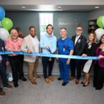 A group of people stand in an office holding a blue ribbon for a ribbon cutting ceremony at AdventHealth Some people are holding scissors, and there are balloons and a plant in the background