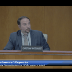 A man in a suit sits at a desk with a microphone, speaking A nameplate reads "Christian Whitehurst " The screen footer says "Commissioners' Reports, Board of County Commissioners, February ,