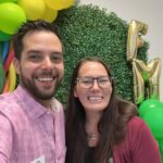 Two people smiling for a photo in front of a green leafy backdrop with yellow balloons and gold letter balloons The man is wearing a pink shirt and name badge; the woman is wearing glasses and a maroon sweater